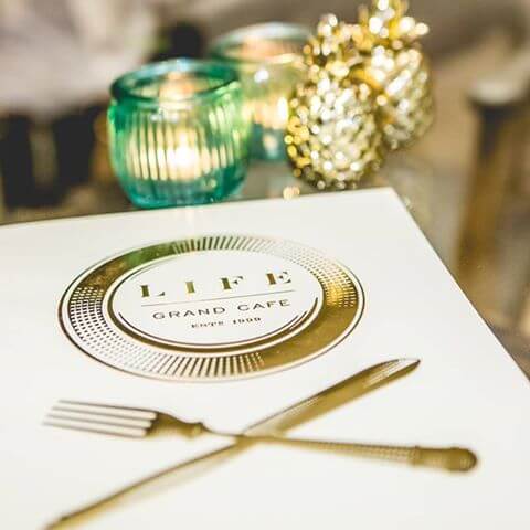 A menu with gold embossed on a white background, a plate encircling the words Life Grand Cafe ESTd 1999, below is a fork and knife crossed. 